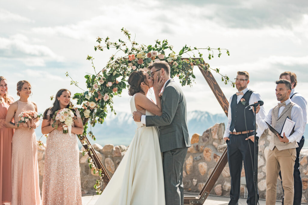 Kayleena and Dominique's first kiss as newlyweds, surrounded by their wedding party and a hexagon arch decorated with florals and greenery at Mt Naomi Vineyards.